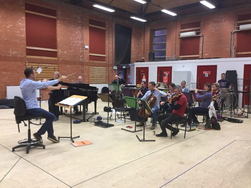 Conductor leads chamber ensemble. He sits on a chair. Musicians play string, woodwind and percussion instruments. Composer in Residence at RSC.