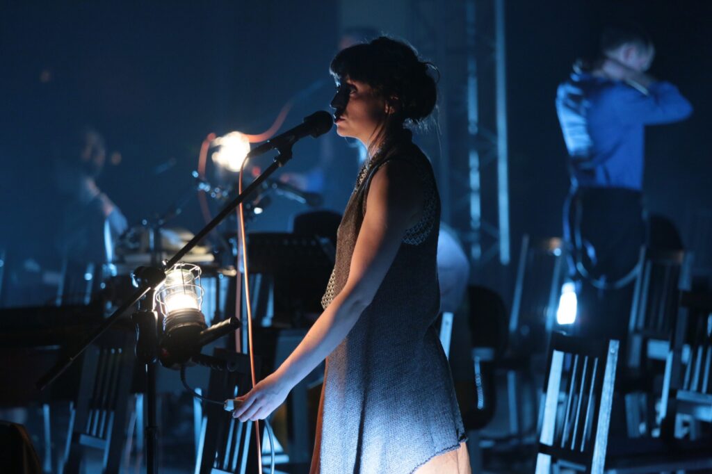 Tale of Two Cities, Chung Ying Theatre production picture. Woman singing original score by Sarah Llewellyn through a microphone in low atmospheric light.