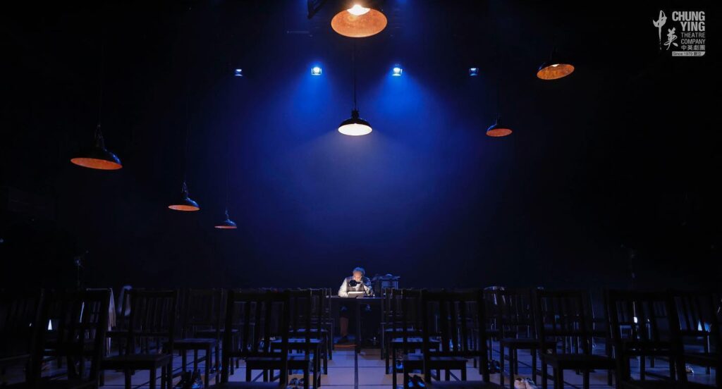Image of performance of A Tale of Two Cities at Chung Ying Theatre. Rows of chairs with actors on stage atmospheric lighting in blue and white.