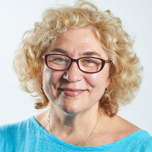 Woman in her 50s smiling with curly blond short hair. Wearing glasses and a blue top. Head shot