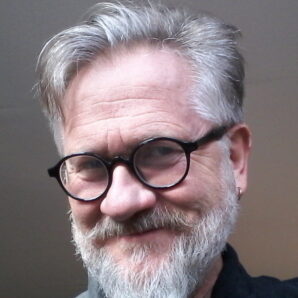 Head shot of theatre director Jonathan Holloway. White man in his 60s with grey hair, moustache and beard. He wears glasses and smiles with mouth closed.