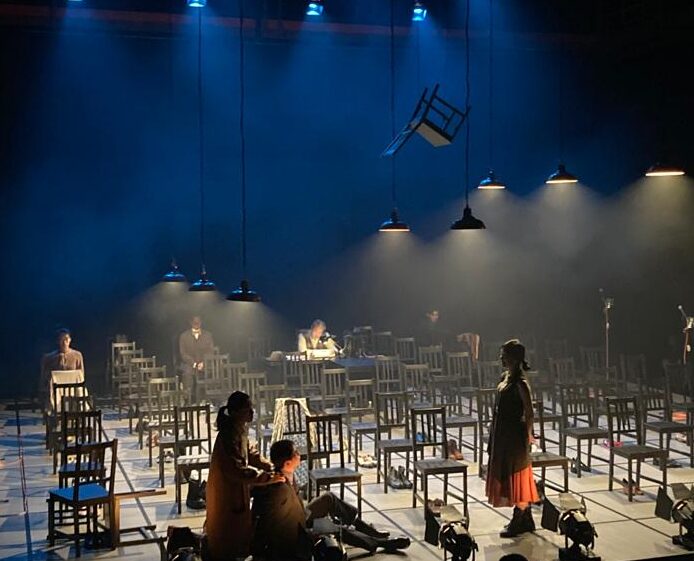 Image of performance of A Tale of Two Cities at Chung Ying Theatre. Rows of chairs with actors on stage atmospheric lighting in blue and white.