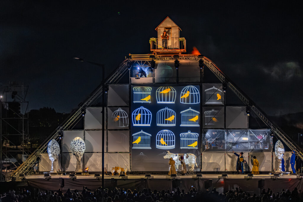 Production image of outdoor theatrical spectacle Ghost Train. 12 meter high stage with a shed atop. Projections of canaries in cages and an actor playing a coal miner looking out of top left corner of stage. Over-sized illuminated cow and trees on stage with actors.