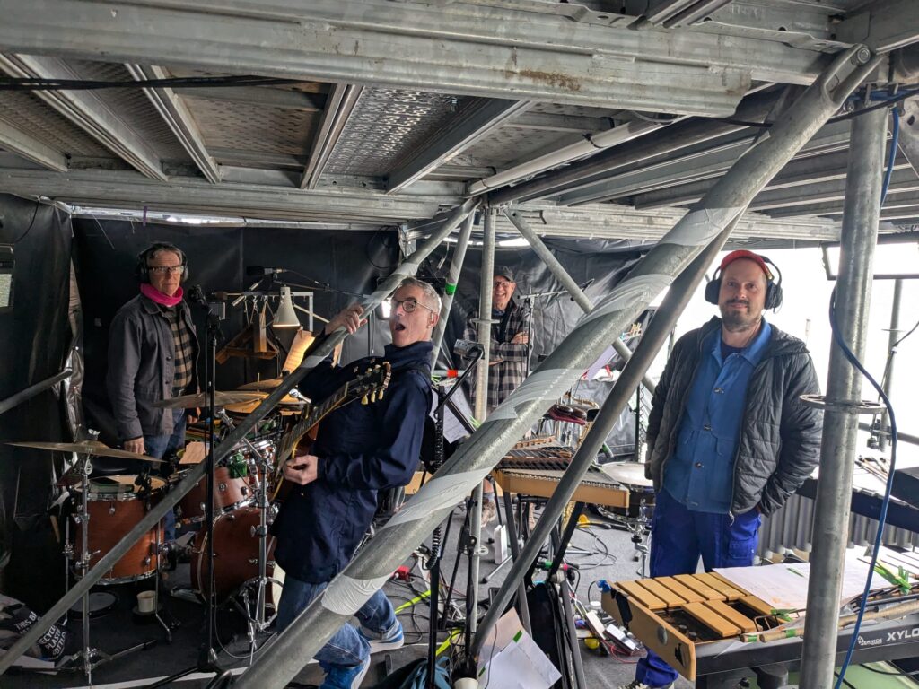 4 male musicians setting up in band box inside scaffold outdoor structure. Percussion instruments and guitar. Faces smiling.