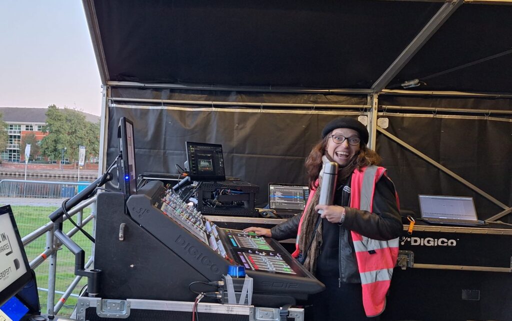Female composer Sarah Llewellyn at mixing desk for large scale outdoor production. Wearing pink hi viz, beret and holding coffee falsk