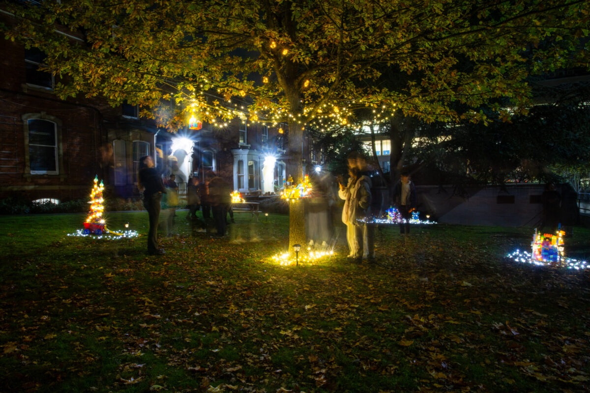 Installation for Leeds Night Light festival. Illuminated fairy houses. One sited around trunk of tree. Two audience members stand next to tree.