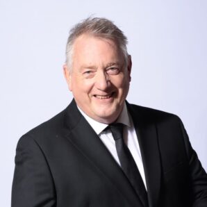 Head shot of white man with grey hair in his 60s smiling. Wearing a black tuxedo and black tie