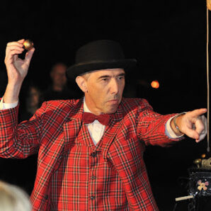 White middle aged man with black top hat, red bow tie and red tartan suit. He holds a gold ball in left hand and points with right hand.
