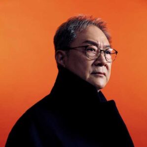 Head shot of Dominic Cheung - Chung Ying Theatre Company. Asian man in his 60s wearing glasses against an orange background.